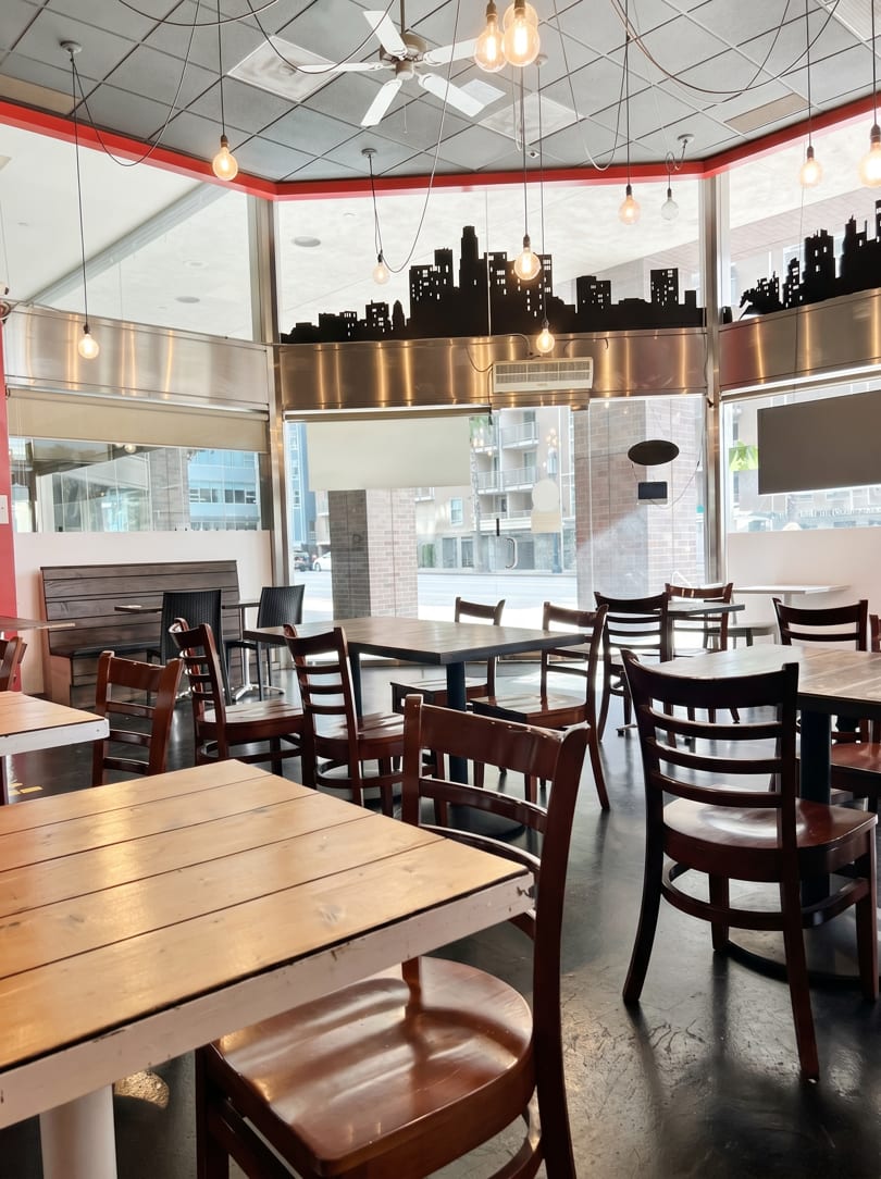 A restaurant dining room with wooden tables and chairs, city skyline decor, and large windows.