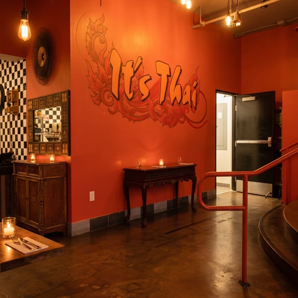 Warmly lit interior of "It's Thai" restaurant with orange walls, decorative bells, and a checkered counter.